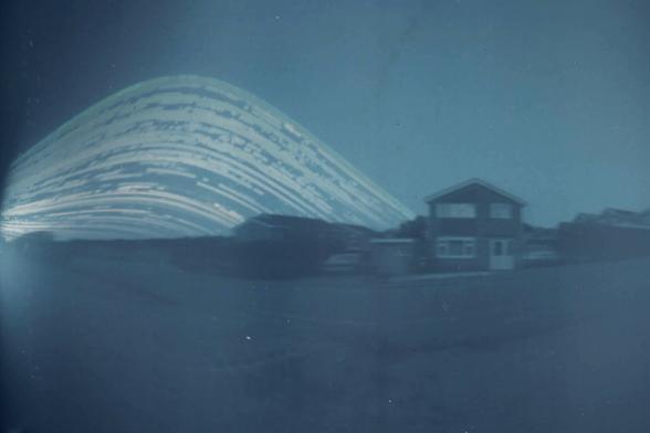 Streaks of light curve across the sky. In the foreground are some houses and a car.