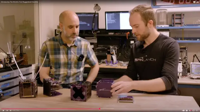 Two people sitting at lab bench looking at cube sats built from purple PCBs