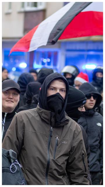 Ein vermummter Neonazi mit Kapuze und Schlauchschal vor seinem Gesicht und dunkelbrauner Jacke.