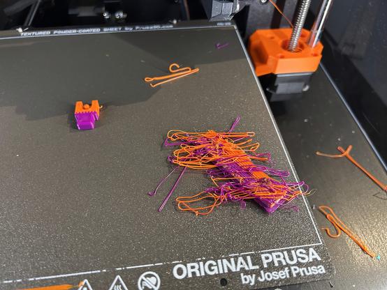 A finished print which is a tiny object half purple and half orange.  It is stuck to the build plate, and is a good print except for one strand of filament sticking out about 3 millimeters from one side.  Also on the build plate is a lot of filament spaghetti and a wipe tower covered in spaghetti filament.  More spaghetti is on the table beside the printer.