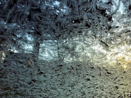 The interior of a car windshield covered with a thin layer of ice in the form of frost ferns. Nothing can be seen beyond the windshield but areas of darkness and light.