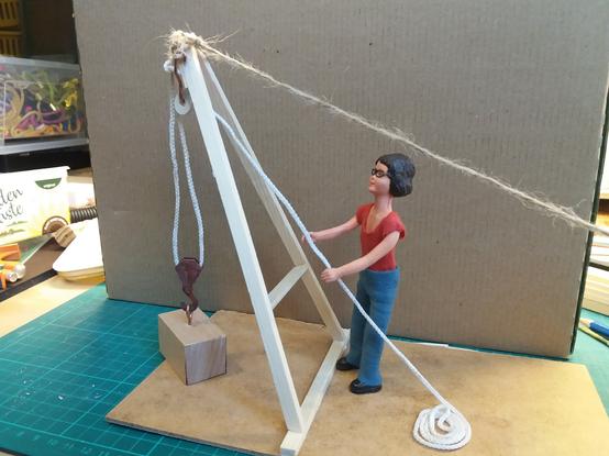 A scale model of a simple crane.
The crane is a simple A frame made from wooden sticks and is about 200mm tall. It is anchored at the base to a small sheet of hardboard, while at the top of the A frame a brown string runs off to the right and is holding the A frame up at a working angle. A pulley is hung from the top of the A frame. A white string runs from the pulley block, down and around another pulley that is hooked to a wooden block, and then back up and over the top pulley. The rope is pulled by Harriet, a 1:12 scale Caucasian doll with dark hair and glasses. She is wearing a red tee shirt and blue jeans.
It was made as a demonstration model for potential museum exhibit.