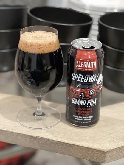 A tall tulip glass of dark beer next to a black pint can. The can has highlights in red and white, a race car and the AleSmith logo are front and center