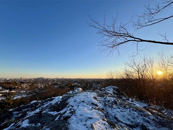 A wide view from the rocky ledge in Middlesex Fells forest: the ledge in the foreground is dusted by snow; the setting sun on the horizon on the right, radiating the tangerine light over the horizon; the Boston skyline is faintly visible on the left; a couple of tree branches in the sky on the right.