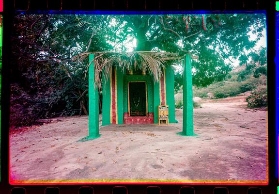 A colorful photo of a bright green, yellow, and red painted small structure, under a tree, housing a small shrine.