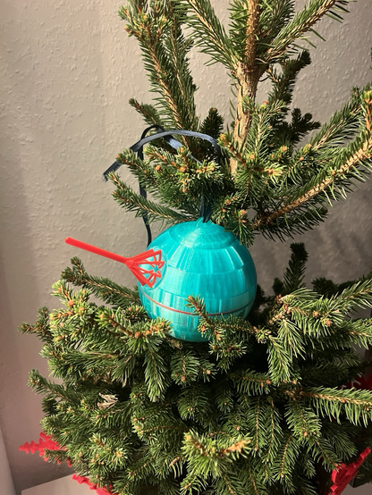 Ein kleiner Weihnachtsbaum mit einem einzigartigen, 3D-gedruckten Weihnachtsschmuck in Form eines Todessterns aus Star Wars. Der Todesstern ist türkisfarben und hat einen roten, detaillierten Laserstrahl. Der Schmuck hängt an einem schwarzen Band und ist gut sichtbar inmitten der grünen Nadeln des Baumes.