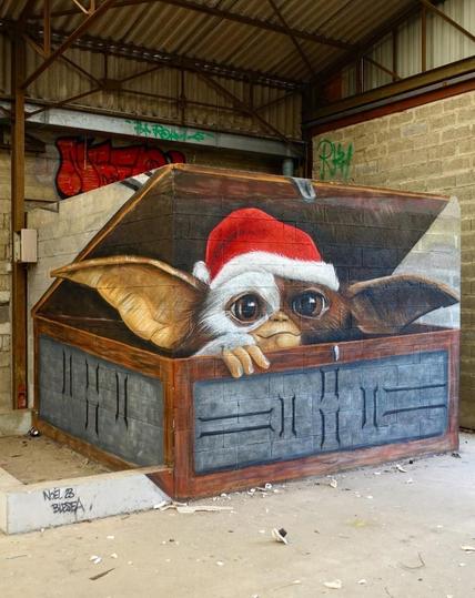 Streetartwall. In an abandoned factory in France, a super cute mural of a Christmas Gremlin has been spray painted/painted in 3D. A square, half-height room (probably an office) is built into a corner of the factory building. The artist has "conjured up" a blue, open wooden treasure chest from which a fluffy creature with long ears and big eyes peers out. It has white and brown fur, a Santa hat on its head and a sad puppy dog look. With one paw on the edge, it probably wants to be let out. 
Info: Gremlins is a 1984 American horror comedy about a young man who keeps a pet mogwai, a small furry imp that, when mistreated, spawns creatures that turn into aggressive monsters and wreak havoc in the city.