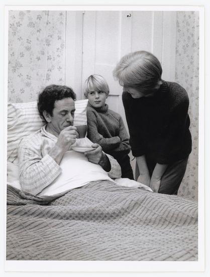 The photograph depicts a moment within what appears to be the domestic setting of a family. In it, an elderly man is seated in bed, sipping from a cup with assistance or presence from two others – possibly his children and wife given their intimate proximity.

On the left side of the image, there's a child sitting on the edge of the bed, dressed in what seems to be casual clothing suitable for cooler temperatures. The middle person appears youthful but not as young as the first individual; they are standing beside him with an attentive posture while holding another cup or bowl.

The adult male is reclined comfortably within his bedroom environment, which includes patterned wallpaper and a floral curtain visible behind them, indicating it could be close to winter time given their attire. The color palette of this photo is monochromatic – black-and-white that captures the scene's emotional gravity without distraction from colors.

This photograph carries an air of quiet domestic life; perhaps capturing a moment during illness or simply everyday familial care. It evokes feelings of empathy, concern for health and wellbeing, and reflects on the roles within family units such as caregiving responsibilities.