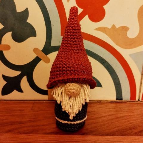 A crochet gnome with a long beard, a big nose and a red pointy hat pulled over his eyes.
