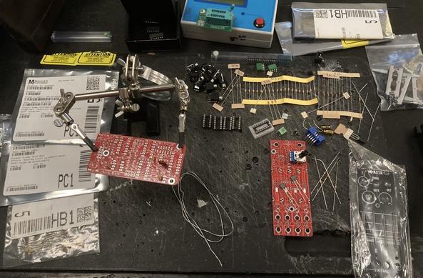 A mess of loose parts to build a Befaco VC ADSR eurorack module, all scattered on a desktop. Resistors, diodes, capacitors, parts in plastic bag. One circuit board is mounted on a helping hand (for soldering). Component measuring tool top center of photo
