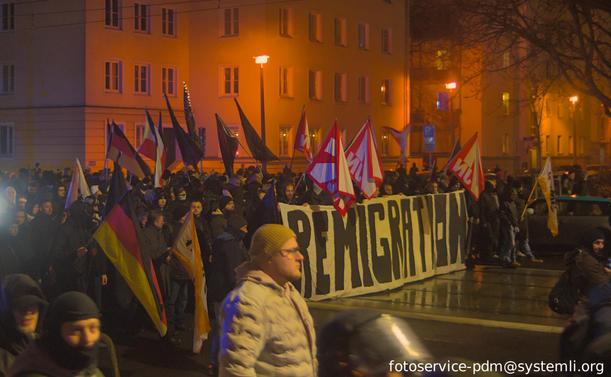 foto vom fronttransparent der rechten demo mit der Aufschrift "remigration", dahinter fahnen der "jungen Nationalisten", deutschland-, sowie reichskriegsfahnen. ein großteil der demo bei nacht ist vermummt