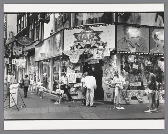 The image is a black and white photograph depicting an urban street scene, likely taken in Amsterdam during the late 1970s to early 1980s. The focal point of the photo centers on "Fashion Stars," a storefront adorned with various posters featuring pop culture icons such as Elvis Presley. A prominent sign bearing the store's name is displayed above the entrance. People are seen engaging in different activities: some browsing items, others conversing, and pedestrians walking by.

The street displays signs for other businesses like "Bambini" and a coffee shop with an arrow pointing towards it. Various advertisements and posters clutter the walls of storefronts and sidewalks, contributing to a bustling atmosphere typical of urban life during that era. The overall impression is one of everyday commerce in a vibrant cityscape.