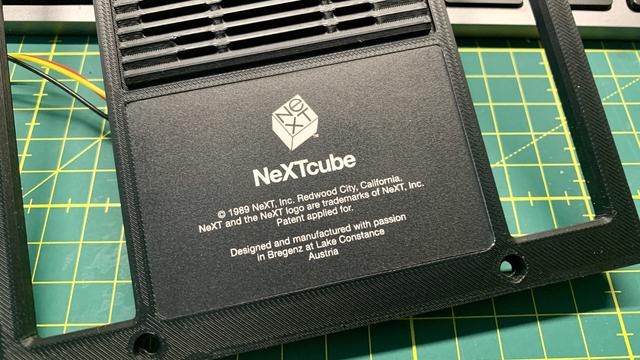 The picture shows the back of a 3D-printed case with the words “NeXTcube” printed on it. Above it is the classic NeXT logo. Below it are the following details:

• © 1989 NeXT, Inc. Redwood City, California.
• Note that NeXT and the logo are trademarks of NeXT, Inc.
• The text concludes with the statement: “Designed and manufactured with passion in Bregenz at Lake Constance, Austria”.

The background shows a cutting mat with markings, and a yellow and a red cable are visible.
