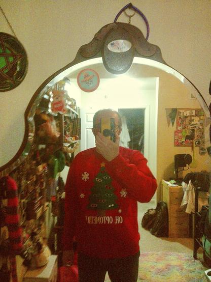 A selfie taken in a mirror. I am wearing a red Christmas sweater with a pattern of snowflakes and Christmas trees on it. Under the tree is the phrase, "Oh, optometree". I am holding a camera up to take the photo. The mirror is ornately framed and has various decorative items hanging around it. The background is a bit cluttered, with shelves, posters, and other objects visible.