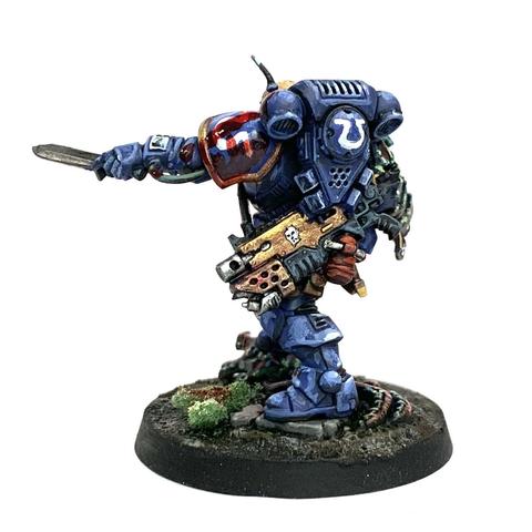 Space Marine in blue power armor with brown pouches belted to him. His right should pad is replaced with some alien chitin plates. He stands on the head of a dead alien with a knife sticking from it. He holds up two more knives, one dripping red gore and the other pointed forward in a challenge. He has blood splattered across his armor.