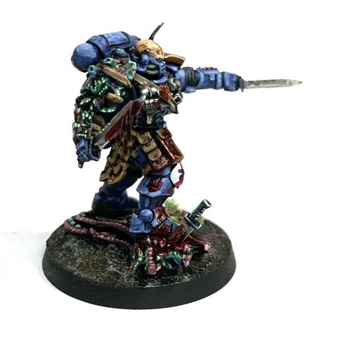 Space Marine in blue power armor with brown pouches belted to him. His right should pad is replaced with some alien chitin plates. He stands on the head of a dead alien with a knife sticking from it. He holds up two more knives, one dripping red gore and the other pointed forward in a challenge. He has blood splattered across his armor.