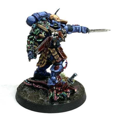 Space Marine in blue power armor with brown pouches belted to him. His right should pad is replaced with some alien chitin plates. He stands on the head of a dead alien with a knife sticking from it. He holds up two more knives, one dripping red gore and the other pointed forward in a challenge. He has blood splattered across his armor.