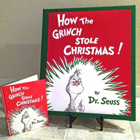 A 2 ft by 3 ft handcrafted version of the original How the Grinch Stole Christmas book by Dr. Seuss (the cover is the one with the pink-eyed Grinch in front of a very spiky white graphic on a red background.) A copy of the original book is in the photo frame for size reference.