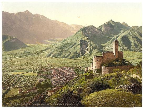 The image depicts a picturesque landscape featuring a historic castle perched atop a hill. The scene is set in the Tyrol region of Austro-Hungary, possibly between 1890 and 1900 based on its vintage appearance.

In this idyllic countryside setting, we observe rolling hills covered with lush greenery that stretches out towards the horizon where majestic mountains rise against a clear sky. The castle stands prominently in the foreground, composed of stone structures exhibiting signs of age and wear. Its architecture suggests medieval design elements such as thick walls and towers for defensive purposes.

Adjacent to the castle is what appears to be an estate with several buildings, possibly including residential quarters or administrative offices, all connected by a network of pathways and roads winding through the property. Vineyards are noticeable in the background, indicating agricultural activity and hinting at the cultivation of wine grapes typical of this region during that era.

The overall ambiance radiates historical charm and serenity, capturing an essence of rural life amid natural splendor with architectural marvels from a bygone period.