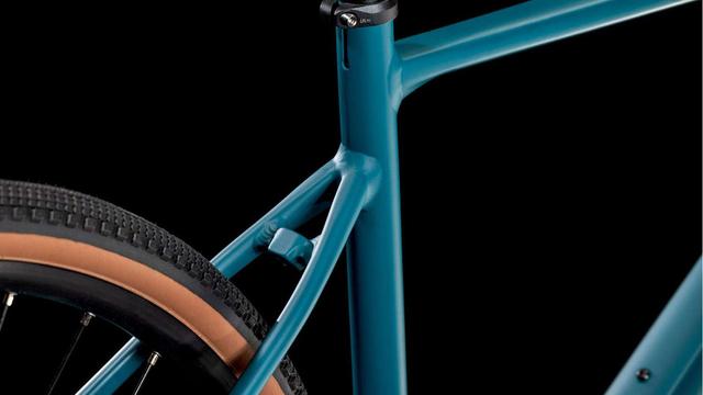 Close-up of a teal bicycle frame focusing on the seat stay area, showing a small, angular metal cube-shaped mount for fenders integrated into the frame design where the seat stays meet. The mount appears as a blocky protrusion against the otherwise smooth frame lines. A gum-wall tire is visible in the background against black.