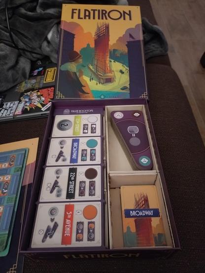 Auf einem Sofa liegen ein Spieledeckel mit dem Schriftzug "Flatiron" und der geöffnete Spielekarton. In dem Karton liegen links die Spieler-Tableaus und Recht sind in zwei Holzkästchen die Ebenenplättchen und Spielkarten untergebracht.