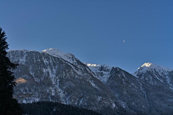 Foto einer verschneiten Bergkette mit Halbmond darüber bei strahlend blauem Himmel.
Links begrenzt ein Thujenbaum / Lebensbaum das Bild, er ist zur Hälfte abgebildet. Verschneite Bäume zieren die Berghänge mit ein paar wenig bewachsenen Rinnen. Die Kette besteht aus 3 Bergen die links in etwa aussehen zwei Höcker mit links dem höheren und Sonnenbeschienenem Gipfel. In der Mitte zieht sich der Gipfel weit nach hinten und ist gerade noch zu sehen bevor er vom Ausläufer des ersten Gipfels verdeckt wird. Der rechte Gipfel sieht aus wie eine Pyramide mit abgeflachter Spitze, die im obersten Bereich sonnenbeschienen ist.