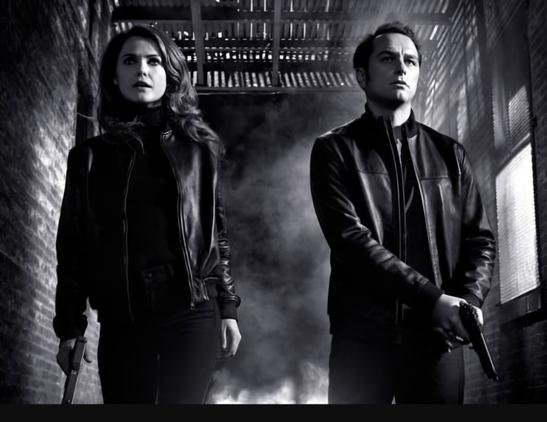 Een zwart wit foto met links Keri Russel als Elizabeth Jennings en rechts Matthew Rhys als Philip Jennings. Allebei in het zwart gekleed en allebei een pistool in de handen. Ze kijken heel grimmig ze staan in een industrieel gebouw met op de achtergrond opstijgende rook.