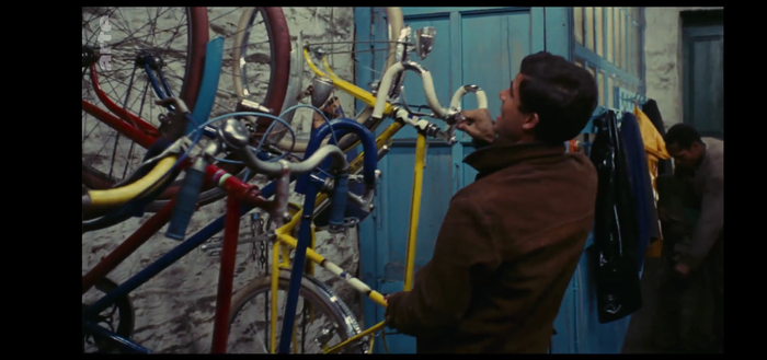 Szene aus dem Film "Die Regenschirme von Cherbourg" von 1963: ein junger Mann nimmt ein französisches Tourenfahrrad von einem Hänger an der Wand. Es hängen weitere Räder dort.