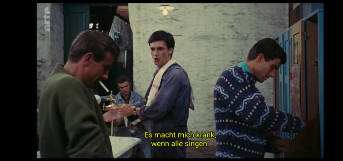 Szene aus dem Film "Die Regenschirme von Cherbourg" von 1963. Männer ziehen sich nach der Arbeit um. Einer sagt im Untertitel: "Es macht mich krank, wenn alle singen."