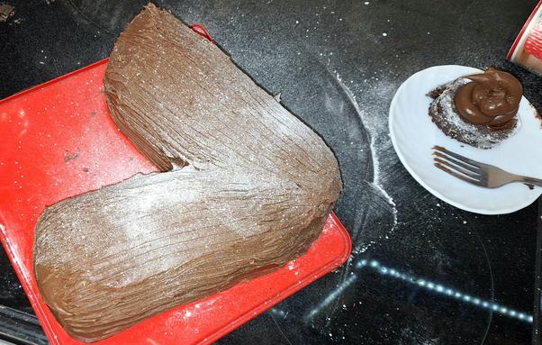 A large, v- shaped cake sits on an upturned Family Circle biscuit box lid. It is brown, lined with bark- like markings and dusted with icing sugar to make it look like a fallen tree at winter. Next to it on a saucer is a spare end with a dollop of ganache by a fork to eat it with. This is all on a black glass cooker top which is also heavily dusted with icing sugar