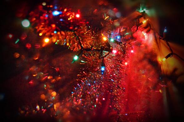 A jumble of light, colour and reflections as a string of small multicoloured lights crosses a couple of tinsel garlands in gold and silver. There is an open net-like fabric with gold thread behind part of it and gold foil paper behind part- we can't really see these in the photo, but they increase the colour and glow and reflections of reflections from the light. The background is generally dark with an overall red glow, black vignette around the edges.