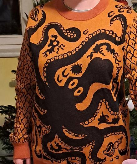 Photo of a sweater. It is orange-brown like pottery, decorated with a replica of the motifs of a Minoan vase in black. In the middle is a huge octopus with large eyes, and around it smaller sea creatures.