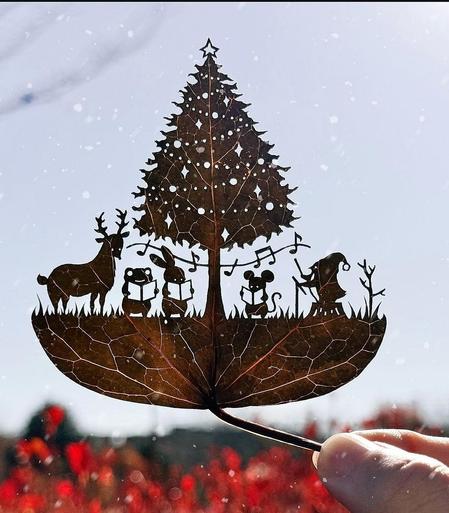 Photography and art. An artfully cut wide leaf as a silhouette with a blurred blue sky and a field of red flowers as a background. The artist uses a cutter knife to cut detailed scenes from leaves. The photo shows a Christmas tree with a star, Christmas baubles and a garland. Below it, five animals stand in the grass and sing a Christmas carol. A deer, a frog, a rabbit and a mouse are holding a Christmas carol in their hands. A little elf with a walking stick conducts the troupe. The filigree work is held by one hand and you can see the blue sky through the cut-outs. Simply beautiful. Merry Christmas.