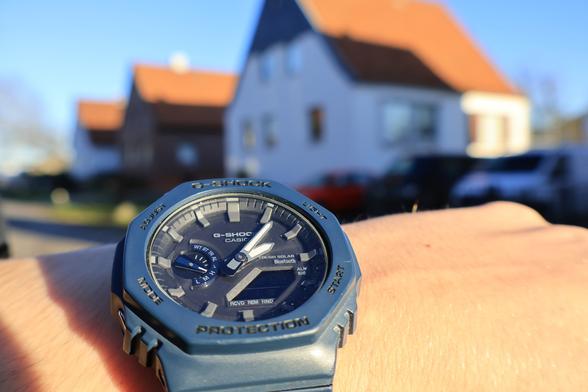 Eine Blaue G-Shock mit analogem Ziffernblat an einem Handgelenk. Im Hintergrund sind unscharf Häuser zu erkennen.