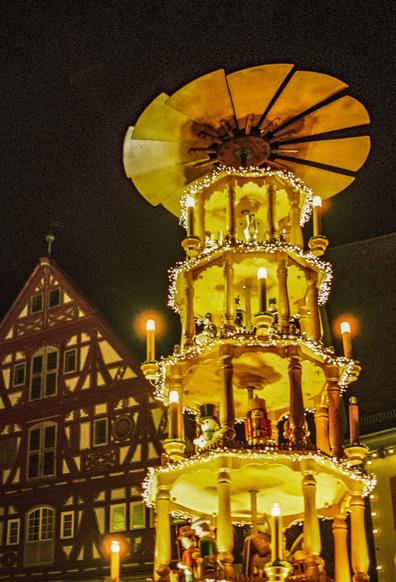 A lit up big wooden christmas pyramide in the right side of the picture frame. In the background in left part of the picture frame we can see a dim lit part of an old city timber-frame house. The sky is dark, showing it has been photographed at night.