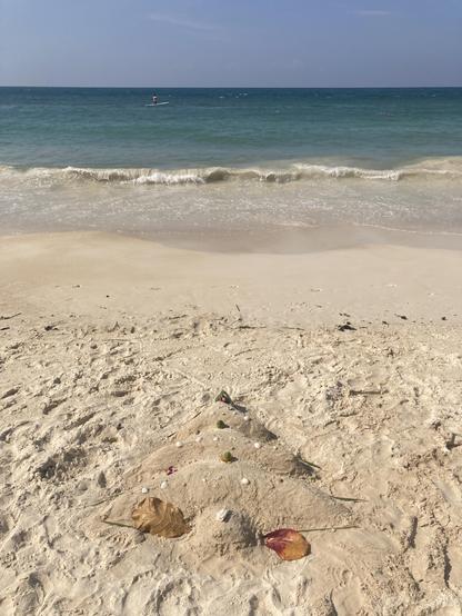 On a white sandy beach a Christmas tree is made out of sand and decorated with some coconuts and leaves. The blue ocean is in the background.