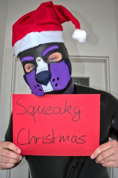 Petplayer in a rubber suit with a Christmas hat, holding a sign saying "Squeaky Christmas"