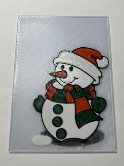 Photo of a 3D printed card on a white background.  The card is transparent with a blue cast.  There is a cartoon snowman wearing a Santa hat, a striped scarf, and mittens.  There is a dark shadow on the ground under him and an unidentified white oval on the ground in front of him.  The snow is ironed white.  The hat brim and tassel are white but not ironed.  The mittens and his buttons are green.  His nose is a carrot, orange.  The scarf alternates red and green and is finished with a Hilbert curve infill.  The shadow is under four layers of transparent filament, so its edges are indistinct.