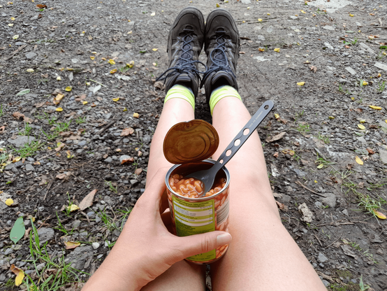 A view of my seemingly bare legs when sitting on the ground. I'm wearing hiking shoes and bright green socks. My hand is holding a can of beans in tomato sauce with a spoon in it.