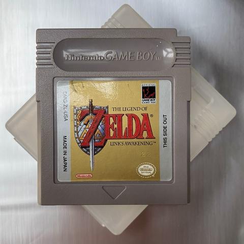 Grey game boy cartridge on silver background with gold label „the legend of Zelda - link‘s awakening“.