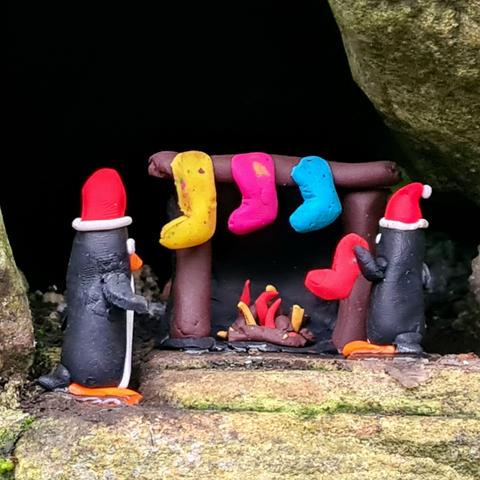 A small model penguin hanging a stocking up on a mantlepeice over a roaring fire.