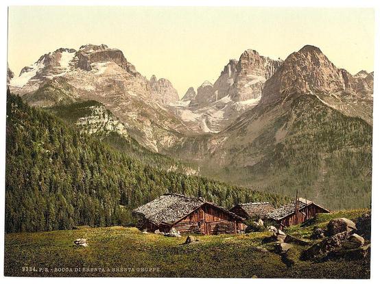 The image depicts a picturesque alpine landscape featuring towering mountain peaks, their rugged cliffs and snow-capped summits rising majestically against the sky. In the foreground, there's a tranquil meadow dotted with scattered rocks, leading up to several traditional wooden chalets nestled among lush greenery. The architecture of these structures suggests they may be used for shelter or leisure by travelers in this mountainous region.

The overall scene is rich in natural beauty and evokes a sense of serenity as it captures the essence of rural life amid such magnificent scenery. It appears to be an early 20th-century photochrome print, given its coloration and style, depicting views from Austria-Hungary's Tyrol during that period.

The photograph serves not only as a visual representation of this stunning alpine environment but also provides insight into the past use of these remote areas by tourists or travelers seeking to immerse themselves in nature.