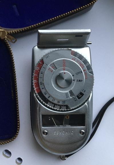 A vintage Sekonic light meter with a silver body and dials for measuring light exposure. It features a circular dial with red indicators for DIN and ASA settings, and a small analog display to show light readings. The background includes a blue case partially visible with a leather outside.