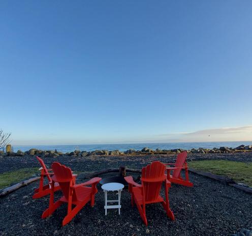 A serene outdoor scene features four bright red Adirondack chairs arranged in a semi-circle around a small white table. The chairs face the calm ocean, with smooth rocks lining the shore in the background. A clear blue sky stretches above, reflecting tranquility and an inviting atmosphere.
