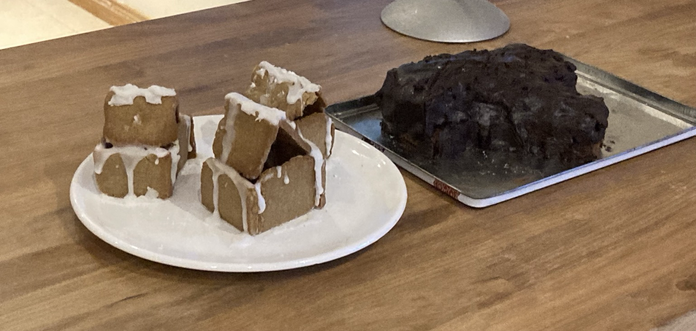 A photo of a plate with three small gingerbread houses with a small amount of white icing on and a chocolate Yule log sitting on a metal lid. Both sit on a wooden worktop.