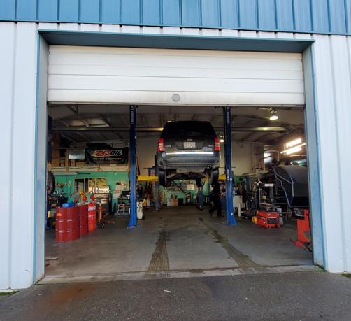 The image captures the interior of a well-lit auto repair shop viewed through an open garage door. A black SUV is suspended on a hydraulic lift, showcasing its undercarriage. The floor is concrete with tire marks leading to the garage entrance. Various tools and equipment can be seen, including red oil barrels on the left and workstations against the green walls. A mechanic, dressed in black, is present, adding to the busy atmosphere of the workshop.