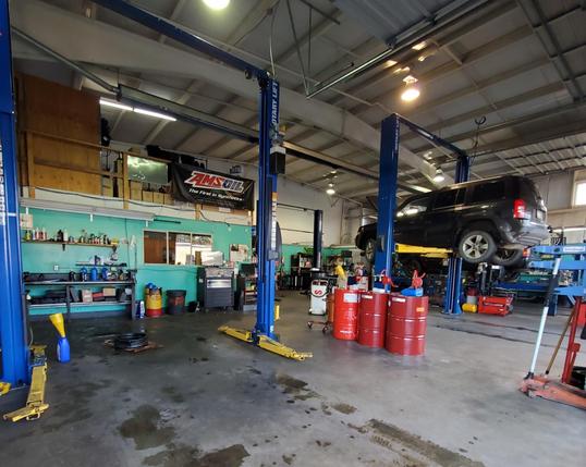 The image depicts the interior of a mechanic's garage, focusing on a black SUV positioned on a yellow hydraulic lift at the center. The ceiling is high with overhead lights illuminating the space. The walls are painted turquoise, contrasting with wooden storage above. On the left, shelves hold a variety of colored containers and tools, while several red barrels are clustered on the right side of the floor. The garage has a concrete floor that appears slightly wet, suggesting recent cleaning or maintenance. The layout is open, allowing for movement around the work area.