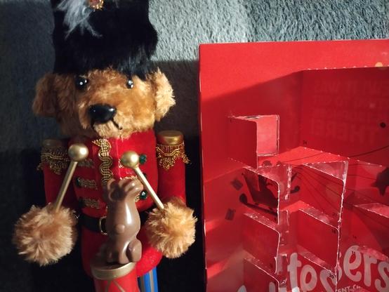 A nutcracker bear in a smart red uniform and playing a drum is standing next to an advent calendar. He has a chocolate balanced on top of the drum.