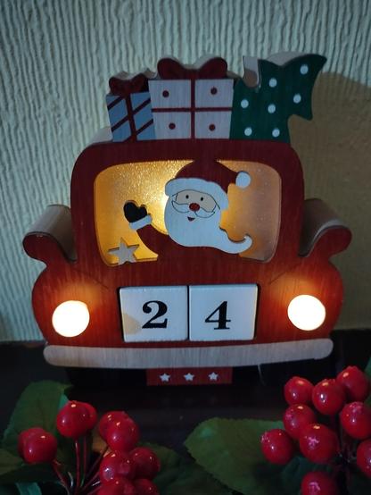 A wooden advent calendar shows Father Christmas driving a red car with presents and a Christmas tree on the roof. The headlights of the car are lit and the number plate is displaying the number twenty four for the twenty fourth day.