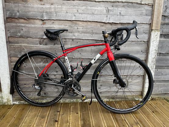A red and black bicycle is leaning against a wooden wall. It has a sleek design, drop handlebars, and two water bottle holders. The bike features fenders and a rear derailleur, resting on a wooden deck.